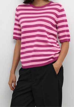 Vrouw draagt een roze gestreepte trui met korte mouwen in licht- en donkerroze, met zwarte broek, één hand in de zak en de andere ontspannen langs het lichaam.