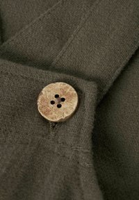 Tessuto verde scuro con una finitura texturizzata, caratterizzato da un pulsante marrone chiaro con quattro fori e un leggero pattern naturale.