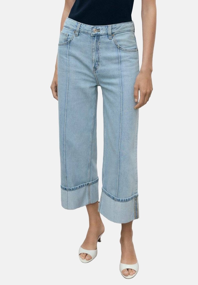 Femme portant un jean large bleu clair raccourci avec des revers, associé à des talons blancs ouverts et un top noir sans manches.