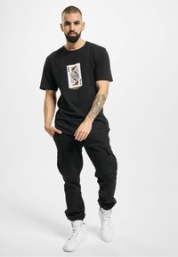 Black cotton t-shirt featuring a colorful playing card graphic on the front. Paired with black cargo pants and white high-top sneakers.