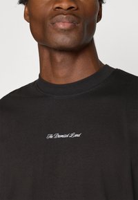 Camiseta preta de algodão com gola redonda, apresentando texto bordado branco "A Terra Prometida" no peito. Textura suave, ajuste casual.