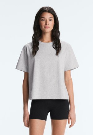 Donna con lunghi capelli scuri, che indossa una maglietta larga grigia chiara a maniche corte e pantaloncini da ciclismo neri aderenti, in piedi davanti a uno sfondo semplice.