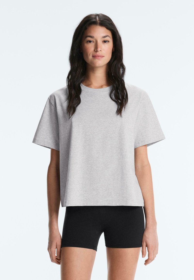 Femme aux longs cheveux foncés portant un t-shirt ample gris clair à manches courtes et un cuissard noir près du corps, debout devant un fond uni.