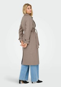 Brun uldblanding trenchcoat med bælte i taljen, slagkrave og slidser i siden. Båret over brede blå jeans og sorte højhælede sko.