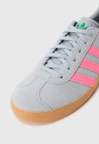 Αθλητικό παπούτσι από ανοιχτό γκρι σουέντ με ροζ ρίγες, υφασμένο λαστιχένιο πέλμα σε φυσικό χρώμα και γκρι κορδόνια. Στο γλώσσα φέρει μικρή πράσινη διακριτική πινελιά με λογότυπο.