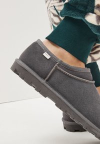Scarpa slip-on in camoscio grigio con superficie testurizzata e profilo basso. Presenta un bordo cucito e un'etichetta del marchio visibile sul lato.