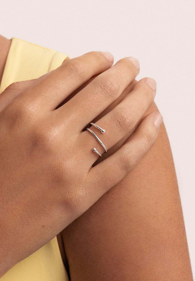 Anillo de plata con dos bandas paralelas y acentos esféricos, llevado en una mano con uñas naturales, mostrando una textura de piel suave.