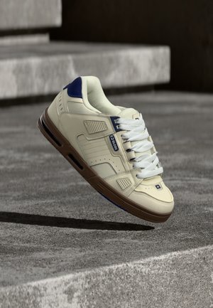 Cream and navy sneaker with brown sole, white laces, and "Globe" logo, hovering above textured concrete steps in shaded outdoor setting.