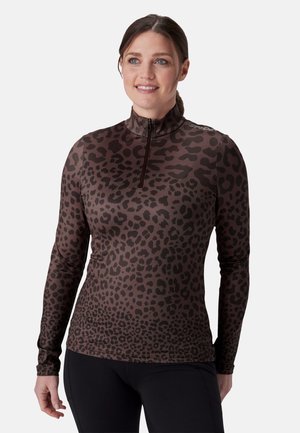Femme aux cheveux attachés porte un haut à manches longues avec demi-fermeture éclair, imprimé léopard marron foncé et noir, et un pantalon noir, souriant légèrement vers la droite.