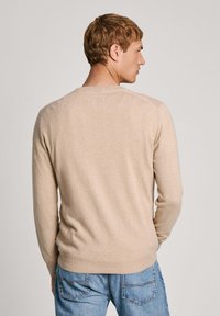 Suéter de punto beige con mangas largas y escote redondo, que presenta puños y bajo acanalados. Llevado sobre unos jeans de mezclilla azules.