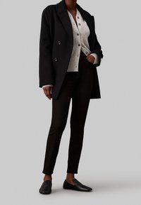 Abrigo negro de doble botonadura, cárdigan gris de punto con botones, pantalones oscuros y zapatos negros tipo slip-on. Tejidos texturizados, corte a medida.