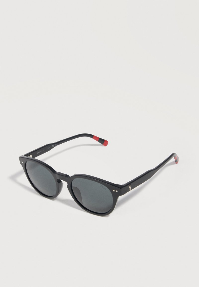 Black round-frame sunglasses with dark lenses and red-tipped arms resting on a light surface with subtle shadows.