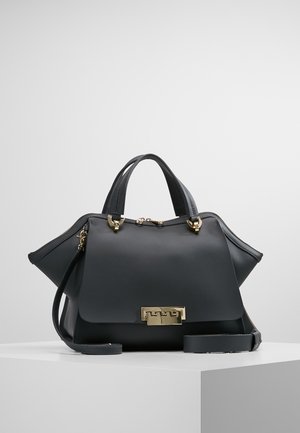 Zwarte leren handtas met gouden sluiting, bovenhandvatten en afneembare schouderriem, tentoongesteld op een wit voetstuk tegen een lichtgrijze achtergrond.