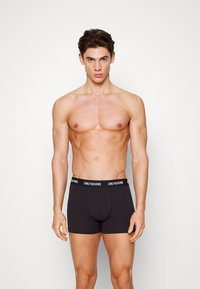 Schwarze Boxershorts aus weicher Baumwolle mit einem logo-bedruckten Bund. Sie bieten eine eng anliegende Passform und eine glatte Textur. Das Model steht vor einem neutralen Hintergrund.