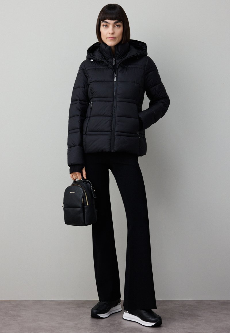 Zwarte donzen jas met capuchon, voorzien van een ritssluiting en zijzakken. Draagt zwarte broek en een kleine zwarte rugzak.