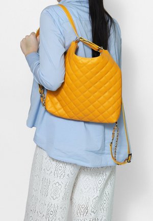 Gepolsterte gelbe Lederhandtasche mit geschwungener Form, goldfarbenen Beschlägen und einem abnehmbaren Kettenriemen, über der Schulter zu einem hellblauen Oberteil getragen.