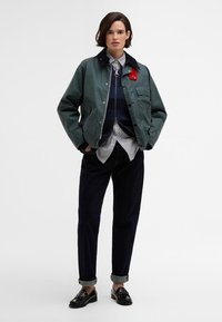 Πράσινη αδιάβροχη ζακέτα με μαύρο γιακά, δύο πλαϊνές τσέπες, φορεμένη πάνω από ένα ναυτικό πουλόβερ, λευκό πουκάμισο και σκούες τζιν. Μαύρα παπούτσια με λεπτομέρειες.