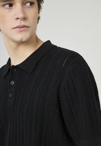 Polo en maille noire avec une texture côtelée, un col et une patte de boutonnage à trois boutons. Motif en relief subtil le long du tissu.