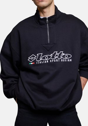 Mann trägt schwarzen Half-Zip-Sweatshirt mit dem Logo "lotto ITALIAN SPORT DESIGN" und kleinem italienischen Flaggenmotiv auf der Vorderseite. Hände in den Taschen.