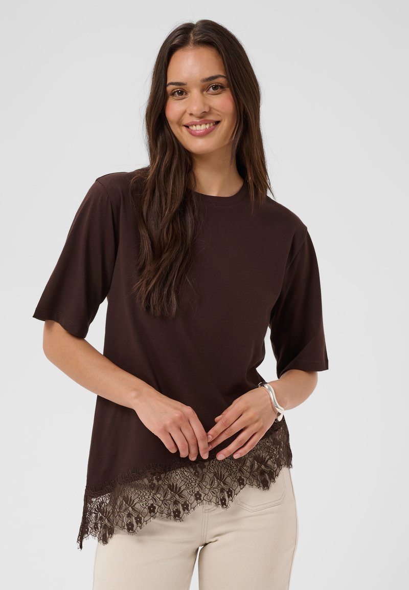 Jeune femme aux longs cheveux bruns portant un haut à manches courtes marron foncé avec bordure en dentelle et un pantalon beige clair, souriant à la caméra.