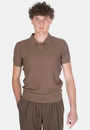 Giovane uomo con capelli ricci che indossa una polo marrone a maniche corte e pantaloni marroni abbinati, in piedi davanti a uno sfondo chiaro e uniforme.