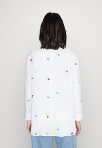 Chemise blanche à manches longues ornée de fleurs brodées multicolores, avec une coupe décontractée et un ourlet droit. Tissu texturé et poignets.