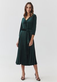 Velvet green dress with a V-neck, three-quarter sleeves, and a pleated skirt. Includes a fabric tie at the waist. Heeled sandals add contrast.