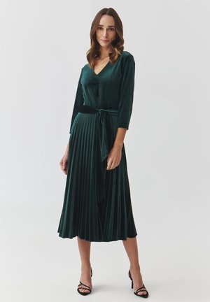 Velvet green dress with a V-neck, three-quarter sleeves, and a pleated skirt. Includes a fabric tie at the waist. Heeled sandals add contrast.