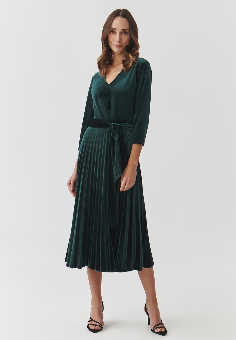 Velvet green dress with a V-neck, three-quarter sleeves, and a pleated skirt. Includes a fabric tie at the waist. Heeled sandals add contrast.