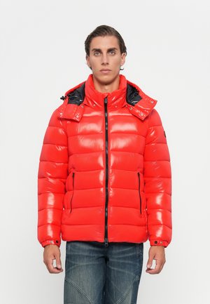 Young man facing forward wearing a shiny red puffer jacket with a hood and dark blue jeans against a plain white background.