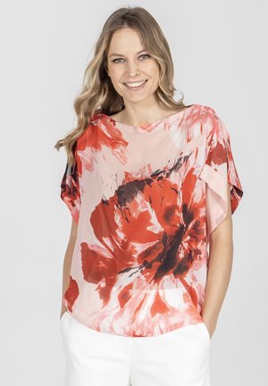 Uśmiechnięta kobieta z falującymi blond włosami nosi białe spodnie i luźną bluzkę z krótkim rękawem z dużym czerwonym, czarnym i różowym kwiatowym wzorem.