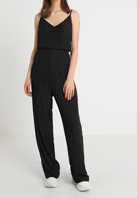 Černý overal s véčkovým výstřihem a tenkými ramínky. Má strukturovanou, žebrovanou látku s akcenty ze stříbrné nitě. Volné kalhoty.
