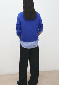 Maglione blu indossato sopra una camicia a righe, abbinato a pantaloni neri larghi. La vista posteriore mostra capelli scuri e lunghi e uno sfondo semplice.