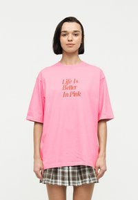 Νέος άνθρωπος με κοντά σκούρα μαλλιά που φοράει ένα φωτεινό ροζ μεγάλος μεγέθους T-shirt με το κείμενο «Η ζωή είναι καλύτερη στο ροζ» και μια καφέ καρώ φούστα.