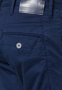 Kalhoty z tkaniny tmavě modré barvy s hladkým povrchem, s zadní kapsou na knoflík a koženou cedulkou s raženým nápisem "RAW".