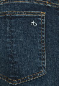 Jeans in denim blu scuro con cuciture arancioni, una tasca posteriore con un logo ricamato bianco e una texture liscia.