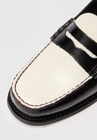 Close-up de um mocassim em couro preto e branco com costura detalhada e fenda para moeda em um fundo branco.