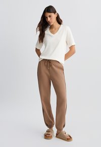 Haut en crème léger avec col en V, coupe décontractée ; pantalon de survêtement marron avec poignets élastiques et cordon de serrage ; sandales beiges à motif tissé.