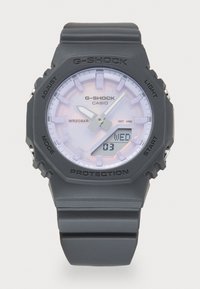 Montre G-Shock avec un boîtier et un bracelet en plastique gris. Présente un cadran rond, un visage de couleur claire avec trois aiguilles, un affichage numérique de la date, et une lunette de protection.