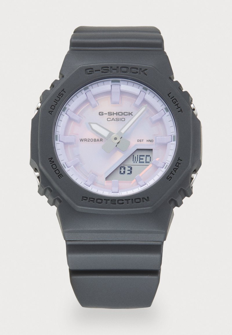 Montre G-Shock avec un boîtier et un bracelet en plastique gris. Présente un cadran rond, un visage de couleur claire avec trois aiguilles, un affichage numérique de la date, et une lunette de protection.