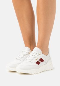 Valkoiset tennarit paksuilla pohjilla, joissa on punaisia ja valkoisia sivuraidoja sekä kultaiset nauhakoukut, käytettynä paljain jaloin valkoista taustaa vasten.