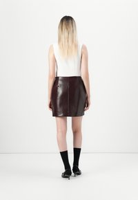Giovane donna con lunghi capelli biondi indossa un top bianco senza maniche, una mini gonna lucida marrone scuro, calzettoni neri e scarpe col tacco nere, in piedi di spalle.
