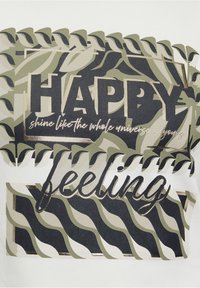 Witte T-shirt met een grafisch ontwerp waarop het woord "HAPPY" in dikke zwarte letters staat en "feeling" in goud, omringd door groene en beige patronen.