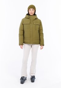 Vrouw in een olijfgroene pufferjack en een beanie, witte snowpants en zwarte skibotten op een witte achtergrond.