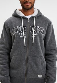 Sudadera gris con cremallera y forro interior blanco, que cuenta con un bolsillo frontal, cordones y el texto grande "CRE EKS ORIGINAL Desde 1974."