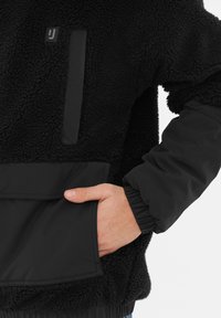 Pullover in pile nero con esterno testurizzato, dettagli in tessuto liscio e una tasca frontale. Le maniche e l'orlo a coste completano il design.
