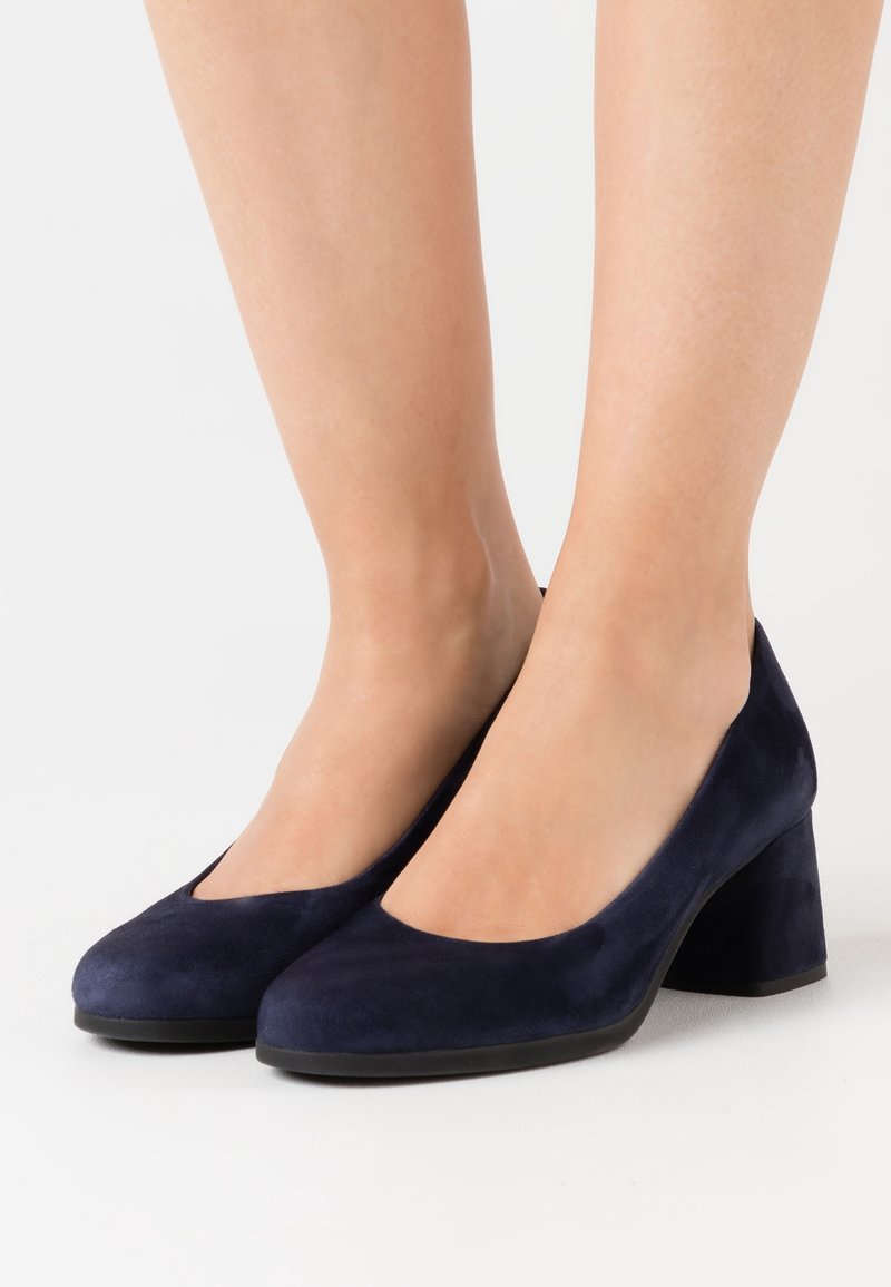 Pieds chaussés de chaussures en daim bleu marine à bout rond et talon bloc sur fond blanc.