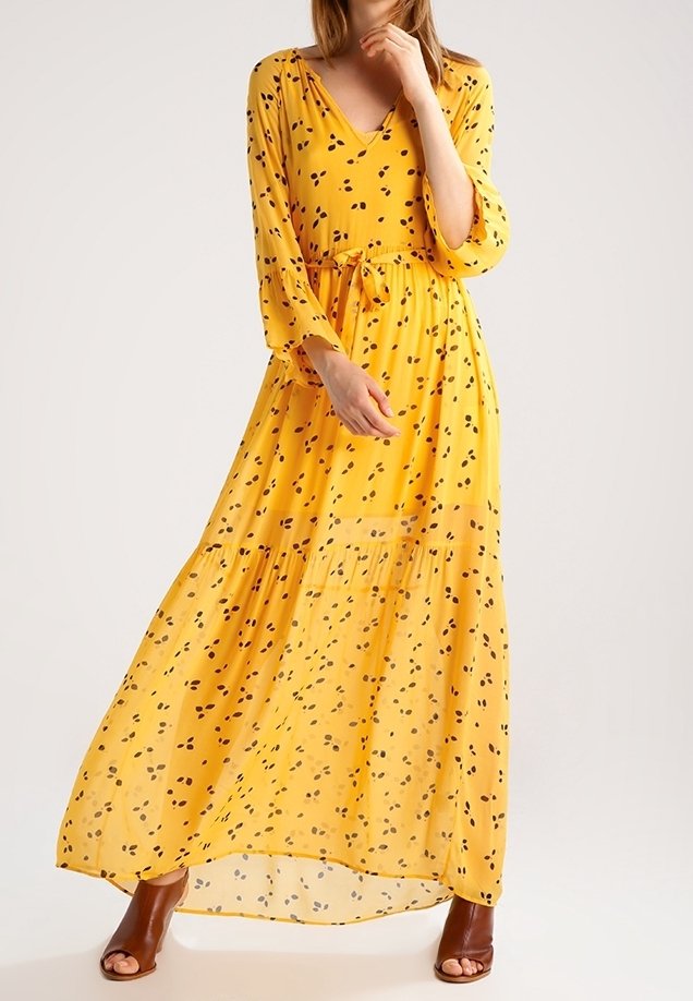 Femme portant une longue robe jaune à pois noirs, manches trois-quarts, et des chaussures à talons ouvertes marron, debout devant un fond uni.