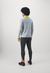 Camiseta atlética de manga larga gris con acentos amarillos, combinada con pantalones atléticos grises con detalles amarillos cerca de los tobillos y calzado negro.