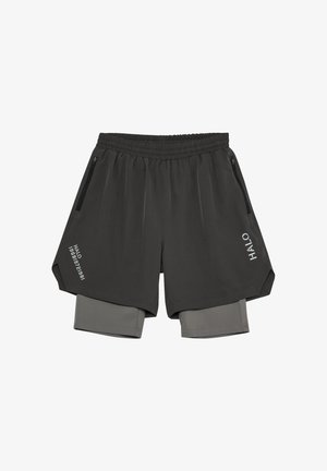 Jonge man staand, gekleed in een grijs mouwloos donsen vest over een grijs kortärmelig shirt, zwarte korte broek, zwarte sokken en zwarte sneakers op een witte achtergrond.